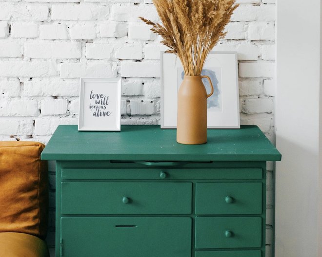 Green wooden dresser with a vase of dried wheat and two framed artworks against a white brick wall